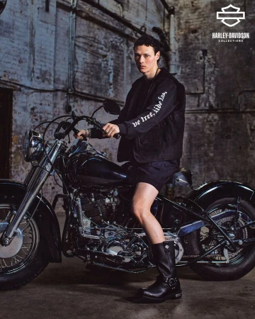 A man in a black jacket sits on a vintage Harley-Davidson motorcycle in an industrial setting. The shot features dramatic lighting and textures.
