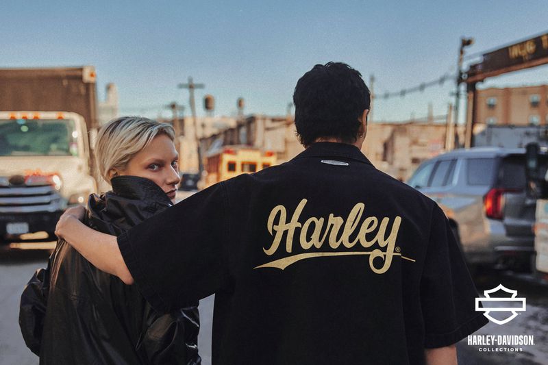 Two people in urban setting, one wearing a black shirt with 'Harley' logo, looking at the camera in an outdoor environment.