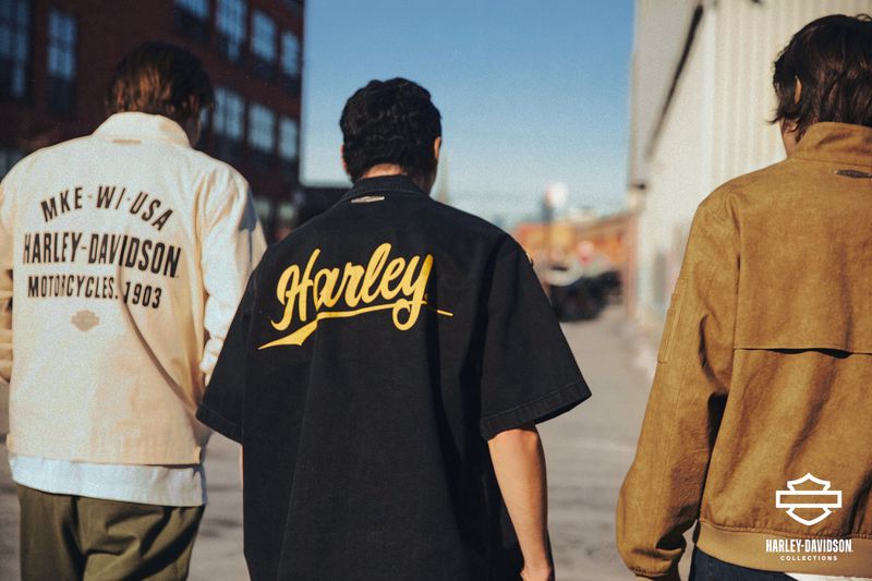 Three adult male models pose from the back in vintage Harley Davidson branded apparel on an urban street with industrial buildings.