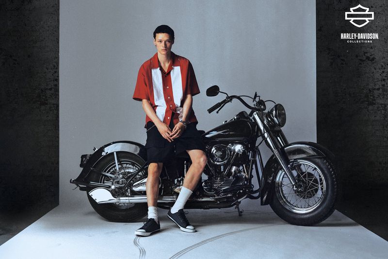 Male model poses next to a vintage Harley-Davidson motorcycle in a studio setting, wearing a red and white shirt and black shorts.