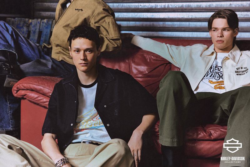 Two young adult men pose on a red couch in a fashion shot featuring Harley-Davidson apparel, denim jackets, and neutral tones.