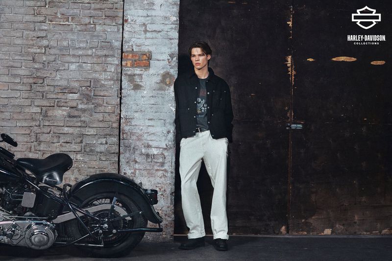 A person standing next to a motorcycle in a vintage garage setting with neutral colors, showcasing fashion and automotive elements.