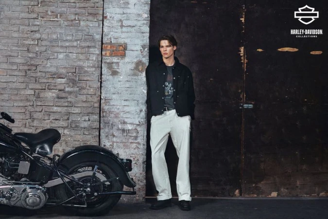 A person standing next to a motorcycle in a vintage garage setting with neutral colors, showcasing fashion and automotive elements.