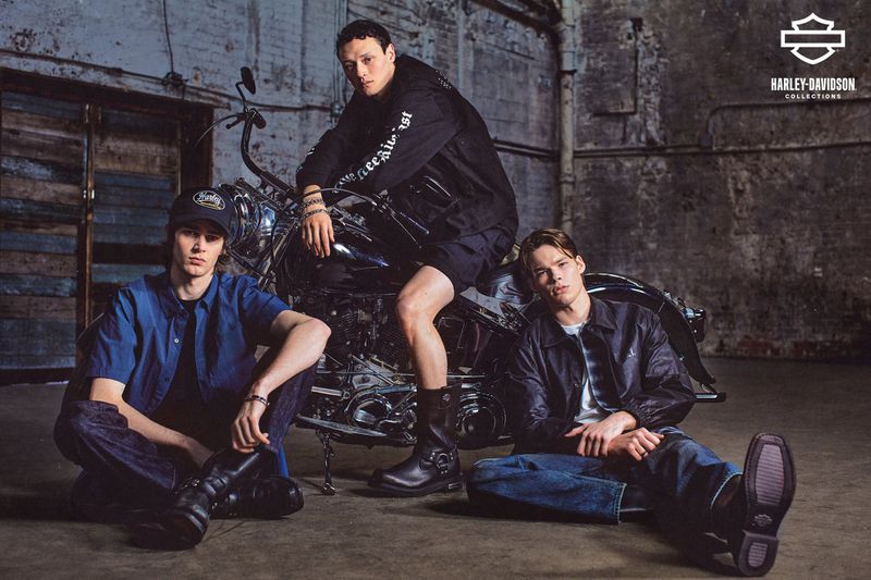 Three adult males showcase Harley-Davidson apparel, seated and posed around a motorcycle in a studio with industrial-style walls and flooring.