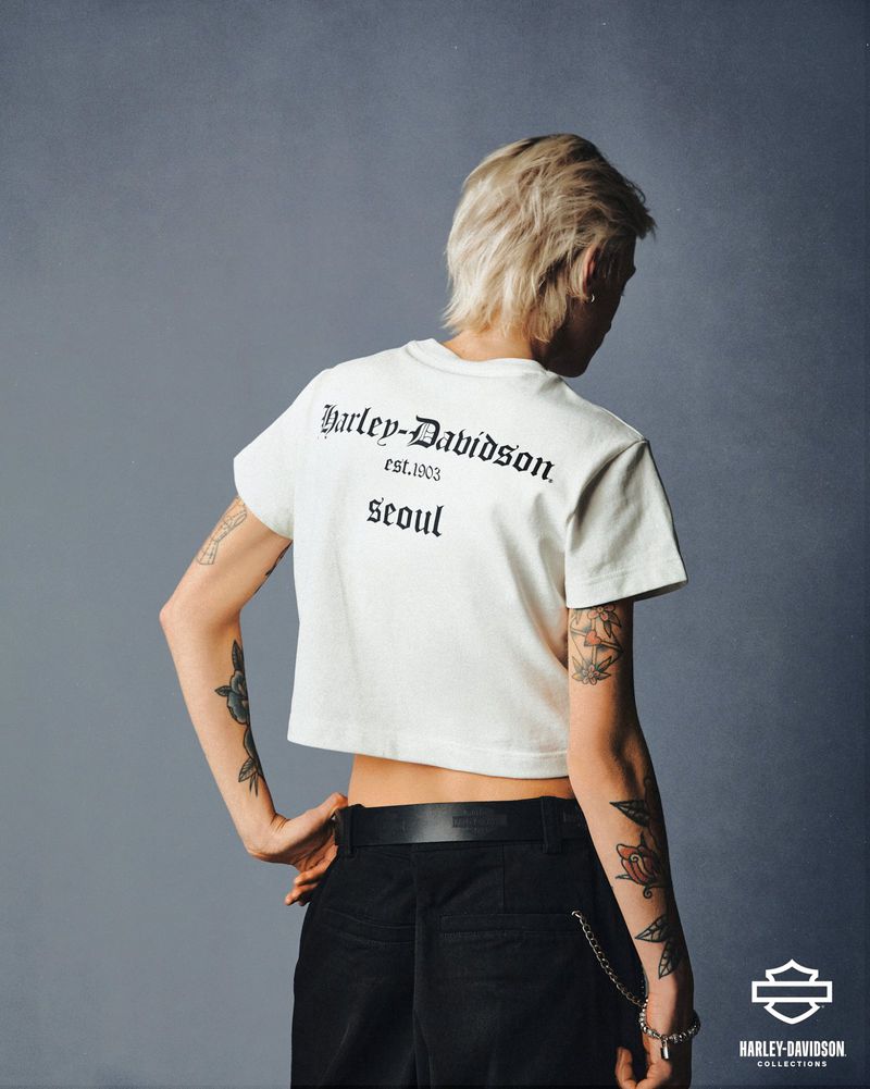 Back view of person in white Harley-Davidson Seoul t-shirt and black pants with tattoos on arms posing against solid grey background.