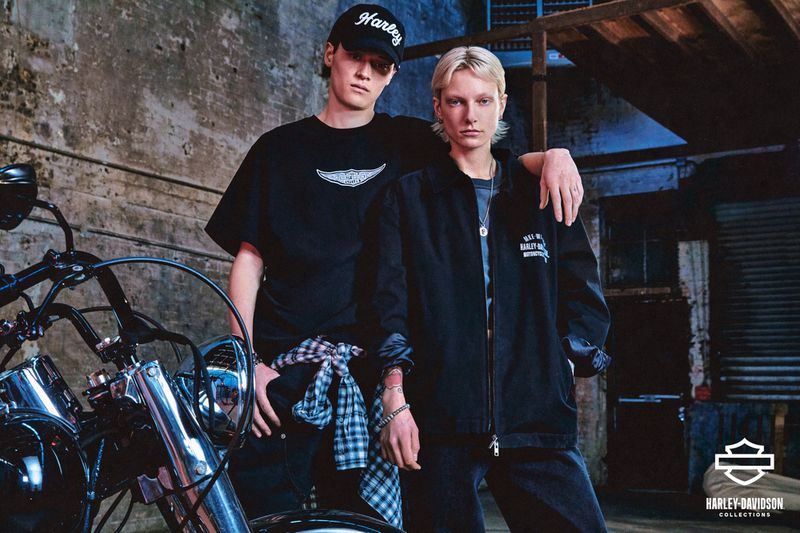 Two people pose with a motorcycle against a textured wall, dressed in Harley-Davidson apparel, under neutral, diffused lighting.