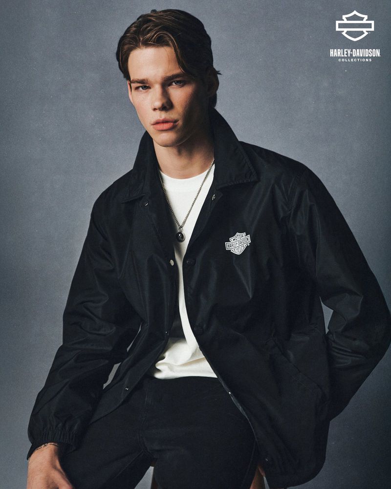 Portrait of a young adult male wearing a black Harley-Davidson jacket and a white t-shirt, posing indoors against a grey backdrop.