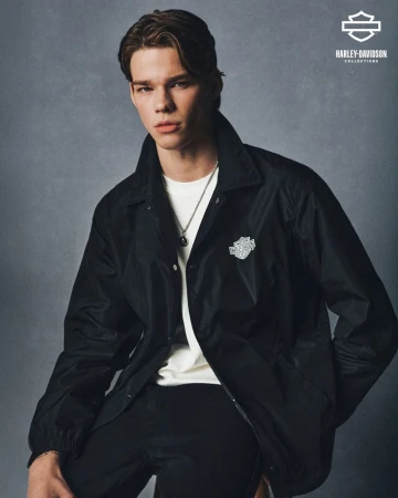 Portrait of a young adult male wearing a black Harley-Davidson jacket and a white t-shirt, posing indoors against a grey backdrop.