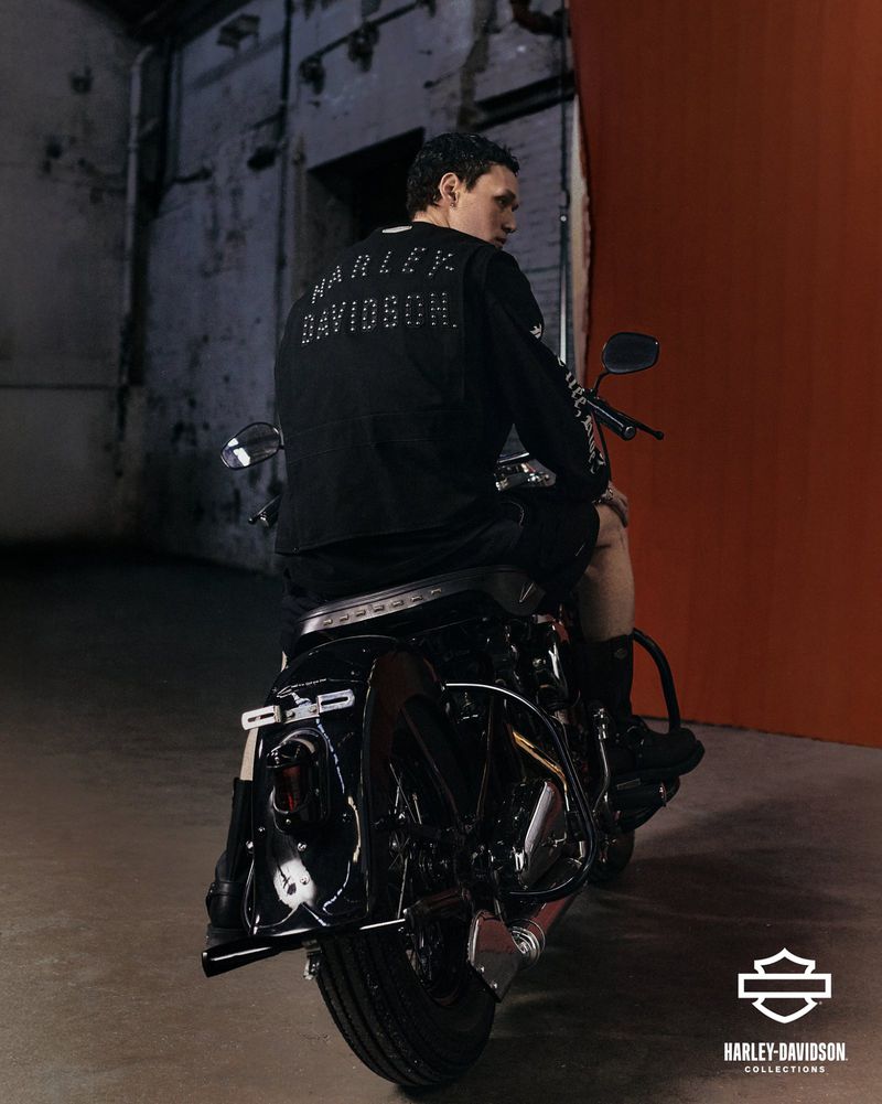 Man seated on a black motorcycle facing away, wearing a black Harley Davidson long sleeve shirt in a concrete garage with orange accent wall.