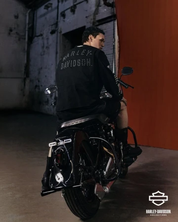 Man seated on a black motorcycle facing away, wearing a black Harley Davidson long sleeve shirt in a concrete garage with orange accent wall.