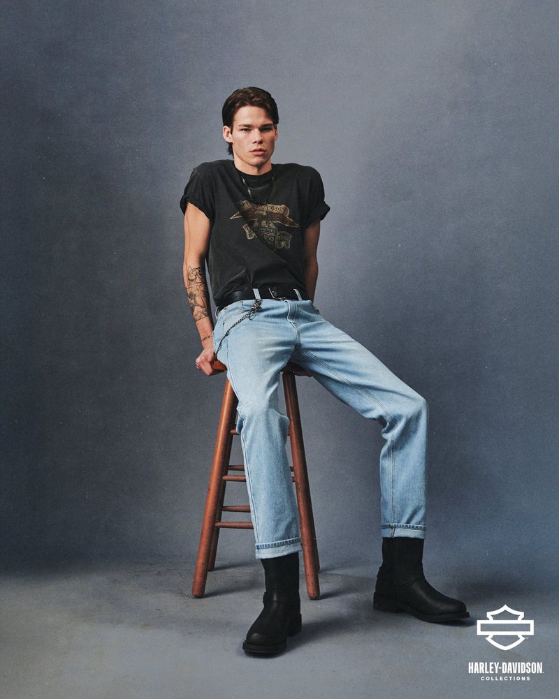 A young adult man wearing a Harley Davidson T-shirt, light-wash jeans, and boots sits on a wooden stool in a studio setting.