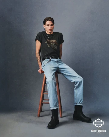 A young adult man wearing a Harley Davidson T-shirt, light-wash jeans, and boots sits on a wooden stool in a studio setting.