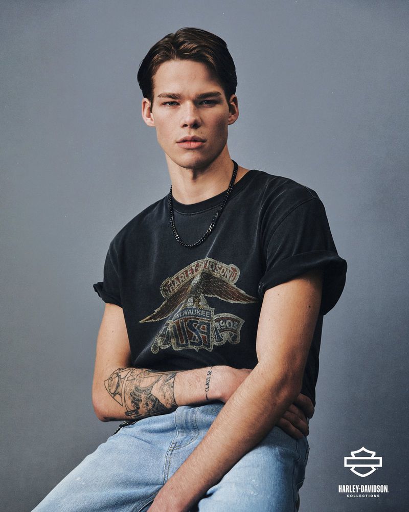 Portrait of a young adult male wearing a Harley-Davidson T-shirt and light blue jeans in front of a plain gray background.