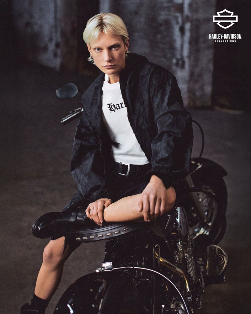 A person with short blonde hair is sitting on a black motorcycle wearing a white t-shirt and black jacket inside a studio environment.
