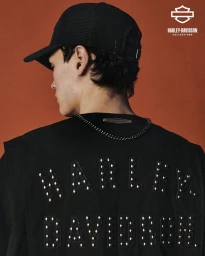 A man wearing a black baseball cap and a studded black vest with Harley Davidson lettering photographed from behind against a rust background.