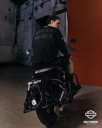 Man sitting backwards on a black motorcycle wearing a black Harley Davidson jacket against a rustic wall background