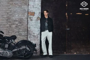 A young male model leaning against a weathered brick wall next to a black motorcycle for Harley Davidson.