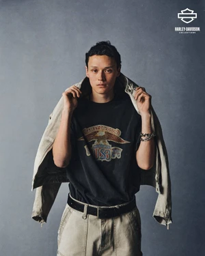 Young man holding light jacket over graphic t-shirt against grey backdrop in Harley Davidson campaign