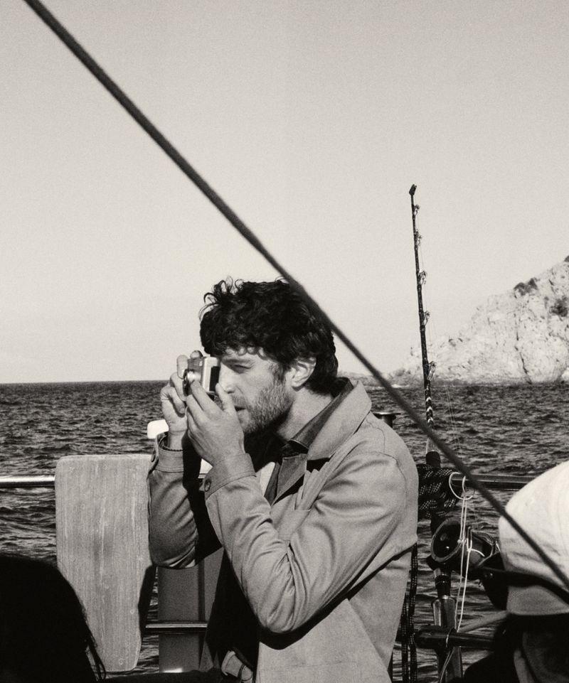 Monochrome medium shot of a man with dark curly hair and a beard holding a camera on a boat with water and land in the background.
