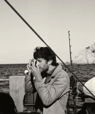 Monochrome medium shot of a man with dark curly hair and a beard holding a camera on a boat with water and land in the background.