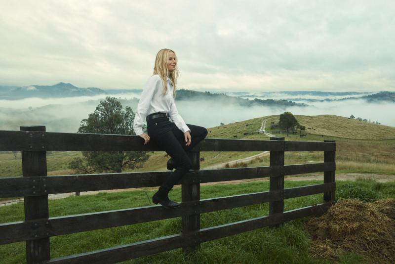 A woman with long blonde hair sits on a wooden fence with a rural landscape of rolling hills and fog in the background.