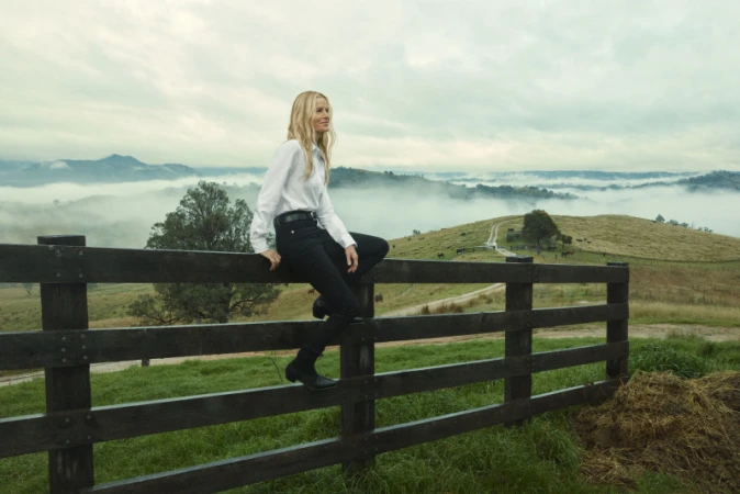A woman with long blonde hair sits on a wooden fence with a rural landscape of rolling hills and fog in the background.