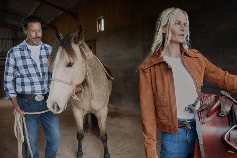 Tan horse with light mane and blonde woman in a brown jacket inside a barn, with a man in a plaid shirt holding a rope.