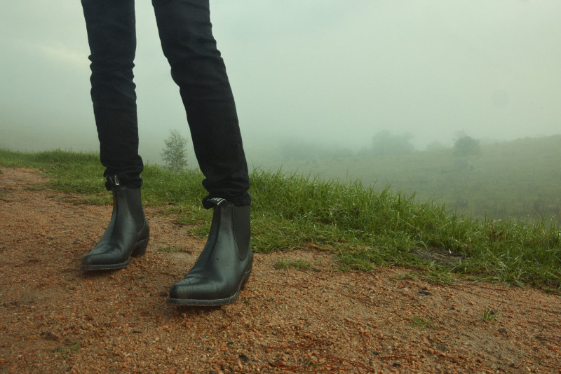 Person stands on a dirt path in black jeans and black boots on a foggy day with green grass and a view of the landscape.