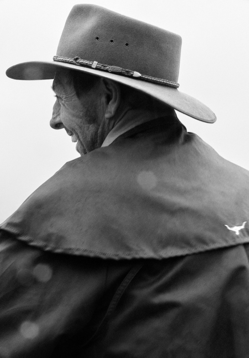 Elderly man with tan skin, wearing a light brown wide-brimmed hat and a dark brown cape, shown in a black and white portrait.