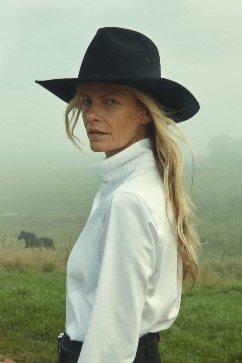 A woman in a cowboy hat and white shirt stands in a field, with a horse visible in the foggy background, in a portrait style.