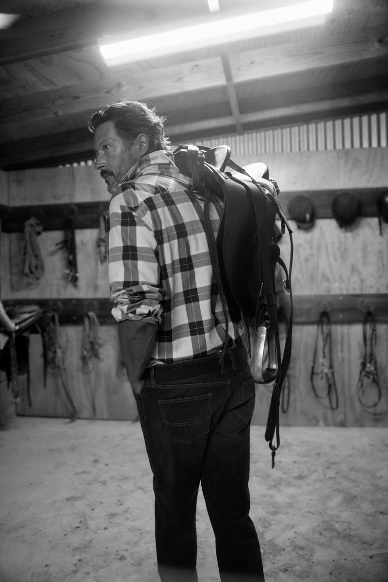 Black and white image of an adult male carrying an English saddle on his back, standing inside a barn with riding equipment.
