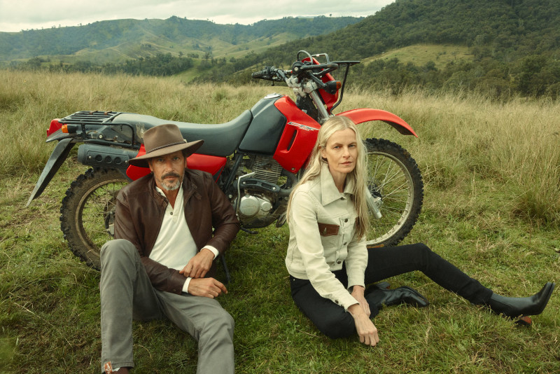 A man and woman sit in a grassy field next to a red and black motorcycle, with rolling hills in the background.