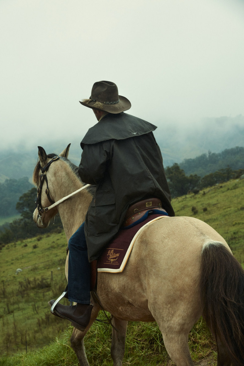 An adult rides a horse through a hilly, grassy landscape, wearing a dark coat and hat, under an overcast sky. Equestrianism theme.