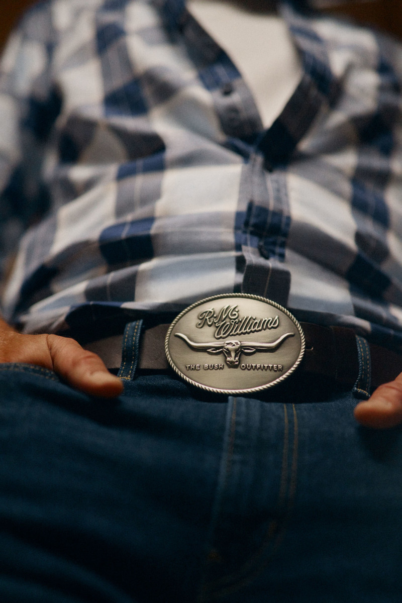 Close-up of torso with plaid shirt, blue jeans, belt buckle featuring longhorns, and weathered hands, creating a rustic western aesthetic.