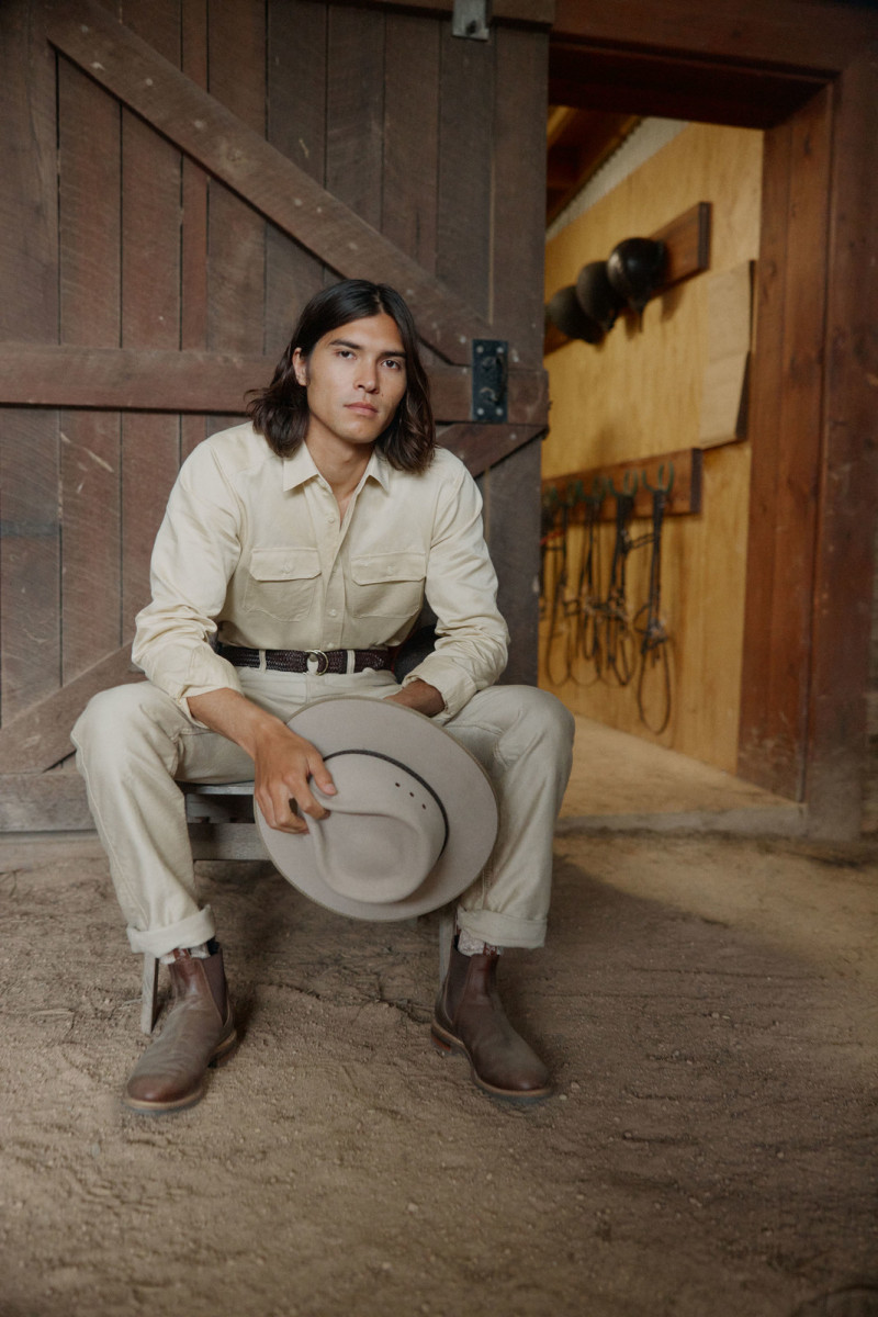 Portrait of a man in a casual outfit sitting in a doorway holding a hat. Rustic wood doors with light brown tones create a rural aesthetic.