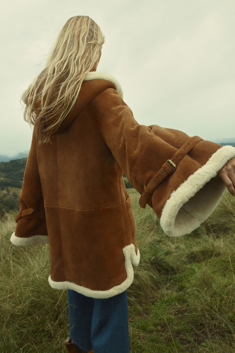 Model wearing a brown shearling coat with cream trim posing in a grassy field with rolling hills under a cloudy sky.