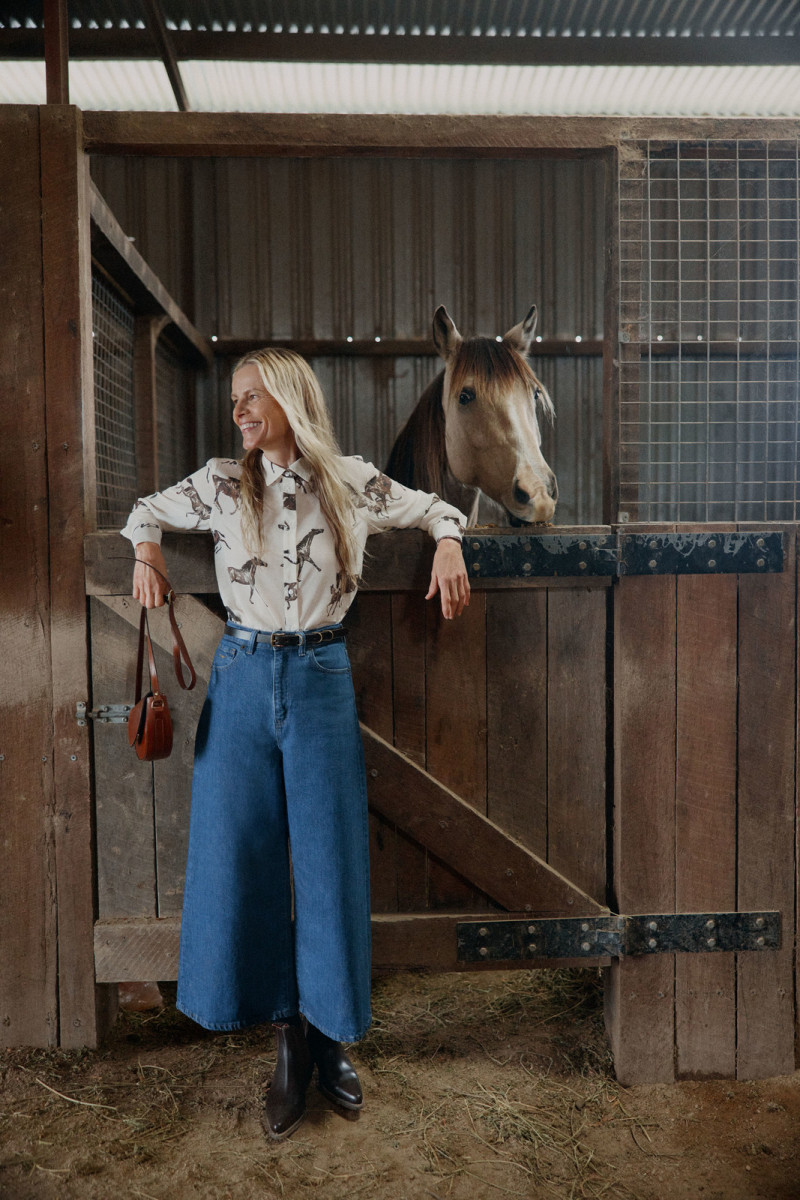 Adult female posing with horse in stable. Woman with long blonde hair wears wide leg jeans and black boots. Horse peeks over stable door.