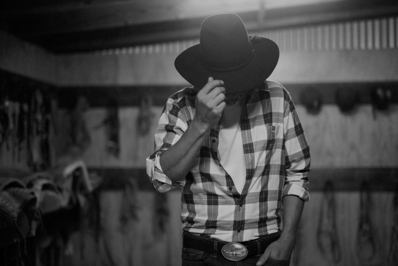 Monochrome shot of an adult male in a plaid shirt and cowboy hat indoors with storage in the background in a medium shot.