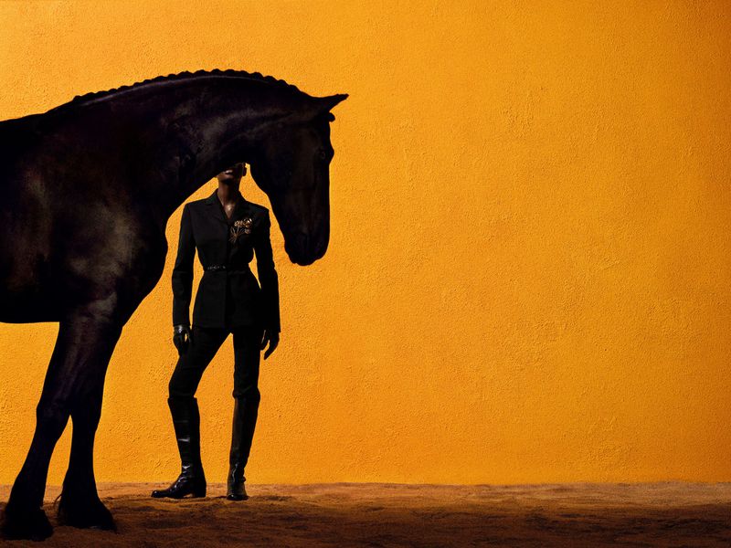 Fashionable woman in dark suit poses with a black horse against an orange background in a studio setting with soft lighting.