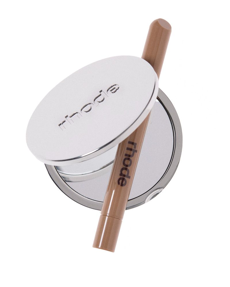 Rhode branded beauty products including a mirrored compact and a lip tint are displayed against a white background in a studio shot.