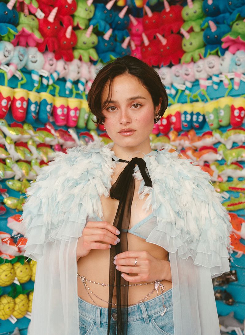 Portrait of a woman with short brown hair wearing a light blue ruffled cape and low-rise jeans, against a colorful plush toy background.