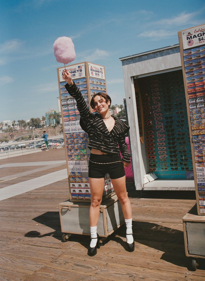 Young woman smiles, holding pink cotton candy near souvenir stands on wooden boardwalk under a blue sky. Fashion shoot.