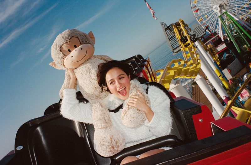 Woman and stuffed monkey toy riding roller coaster at amusement park under blue sky, Ferris wheel and American flag in background.