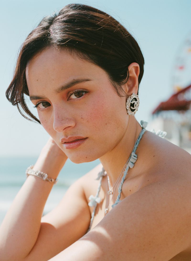 Close-up of a young woman with dark hair and earrings, posing on a beach with the ocean and an amusement park Ferris wheel in the background.