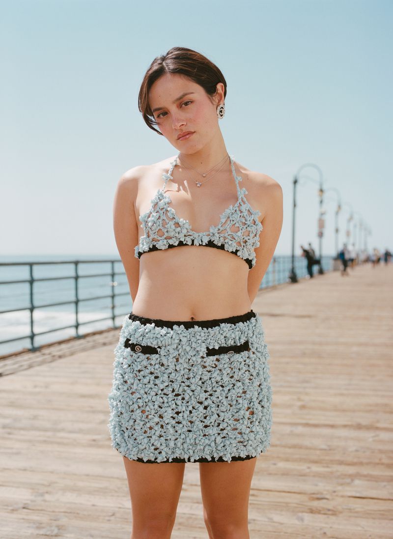 Young woman poses on pier in a light blue crochet two-piece outfit against a backdrop of blue sky and ocean on a sunny day.