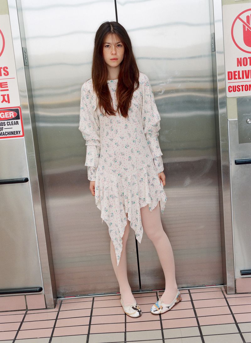 Young woman poses in a floral dress against a metal elevator with tiled floors. Fashion editorial portrait with indoor studio lighting.