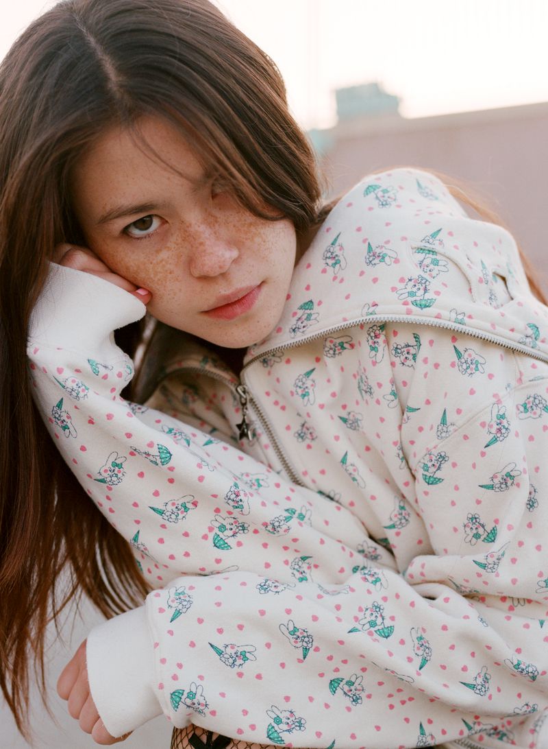 Young woman with freckles in patterned zip-up hoodie poses against a city backdrop, gazing at the camera with neutral expression.