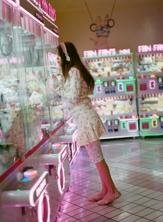 Young woman in floral outfit plays claw machine at an indoor arcade with pink neon lights, creating a whimsical and playful scene.