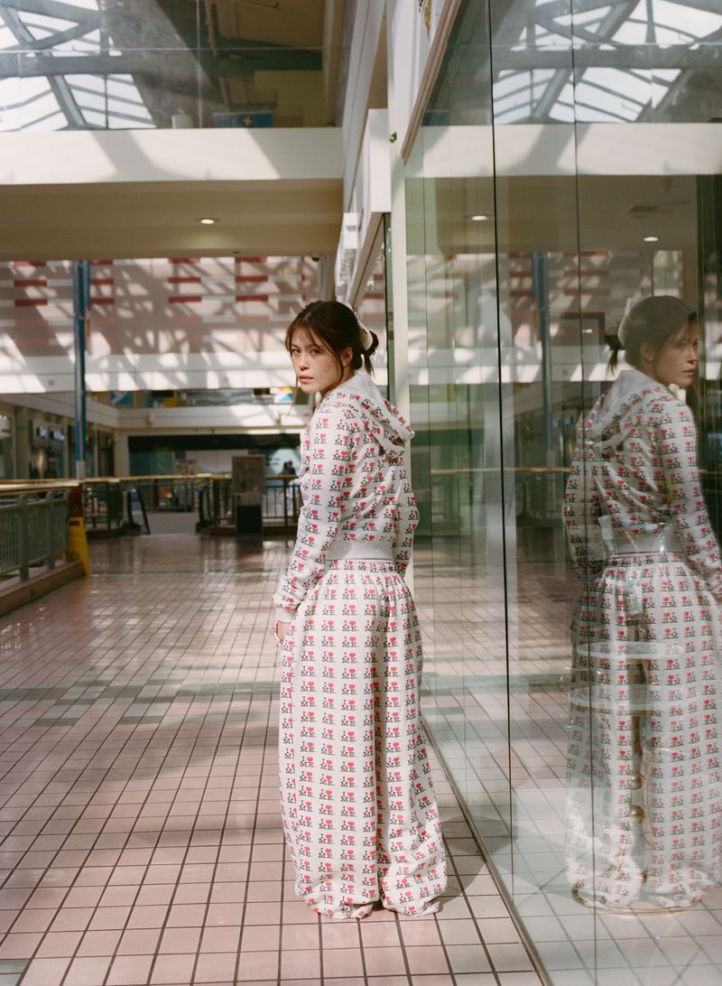 Young woman in patterned white sweatsuit with hoodie looks over shoulder in a glass-walled hallway with a light-tiled floor.