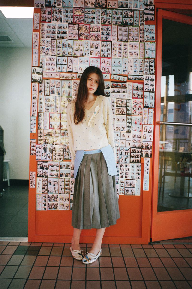 A young woman in a cream sweater and grey skirt stands in front of a wall covered in photos, with a red door frame surrounding her.